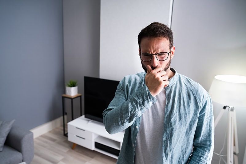 A man with a beard and glasses in a living room setting, holding his hand over his nose and mouth with a disgusted or uncomfortable grimace.